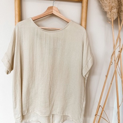 T-shirt bege pendurada em cabide numa escada de bambu com plantas secas ao lado.