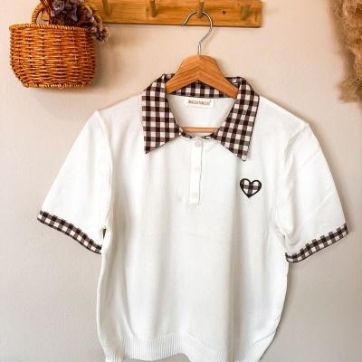 Camisola branca com gola e detalhes xadrez castanho e branco pendurada numa parede clara