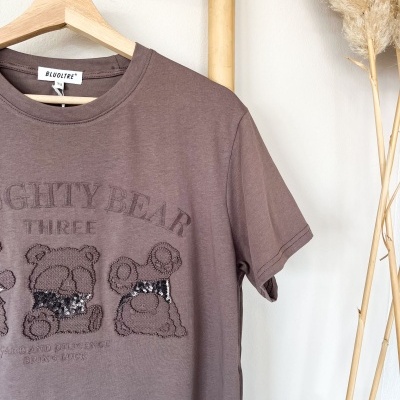 T-shirt castanha com urso bordado e paillettes pendurada num cabide de madeira