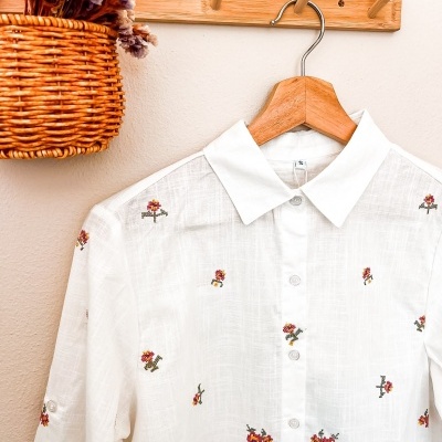 Camisa branca com pequenas flores bordadas pendurada num cabide de madeira e cabideiro de madeira com cesto de vime e flores secas