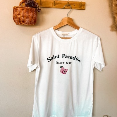 T-shirt branca de algodão com texto e bordado, pendurada em cabide de madeira numa parede bege
