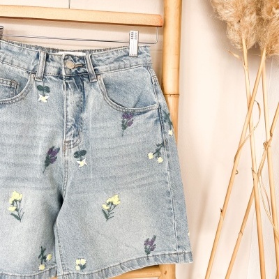 Shorts de ganga com bordados florais pendurados em cabide de madeira com suporte de bambu