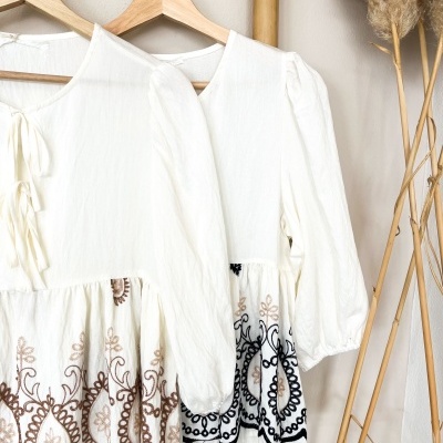 Dupla de blusas brancas com bordados florais castanhos e pretos penduradas em cabides de madeira.