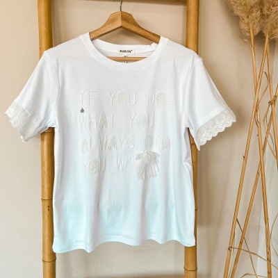 T-shirt branca com mensagem bordada e renda nas mangas pendurada em cabide de madeira.