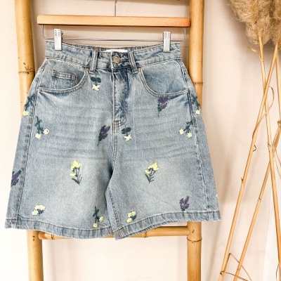 Calções de ganga azul claro com flores bordadas pendurados em cabide numa estrutura de bambu