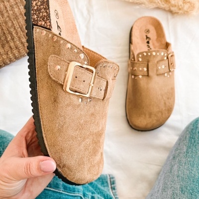 Sandálias castanhas de camurça com fivela dourada e tachas, com sola preta, sobre fundo branco e jeans azul