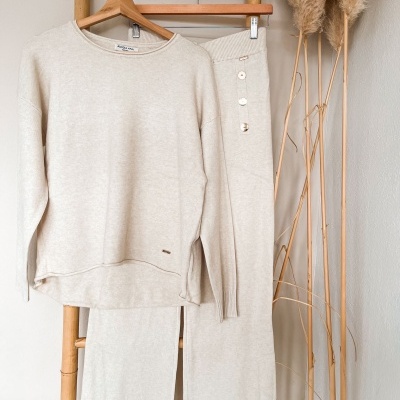 Conjunto de camisola e calças bege claro pendurados em cabides numa estrutura de bambu.