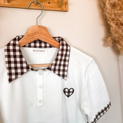 Camisola branca com gola xadrez castanha e branca pendurada num cabide de madeira.
