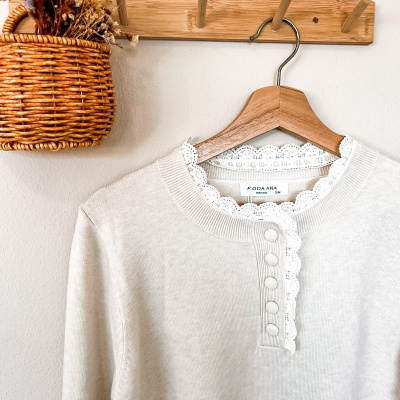 Camisola creme com renda branca no colarinho pendurada em cabide de madeira, junto a cesto de vime com flores secas