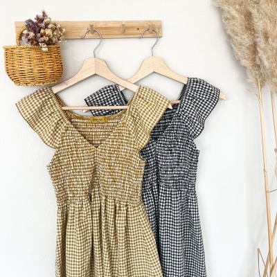 Vestidos vichy castanho e preto pendurados em cabides numa parede branca com cesto de vime com flores secas