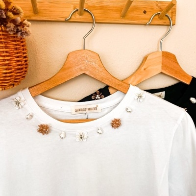 T-shirts branca e preta com detalhes bordados penduradas em cabides de madeira num suporte de parede de madeira
