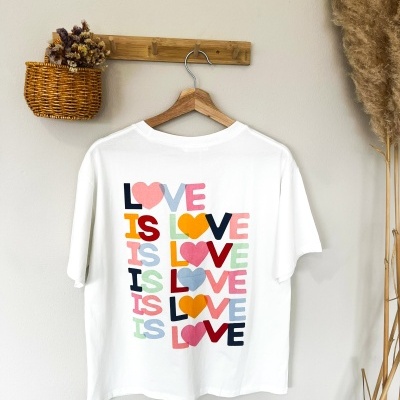 T-shirt branca com estampa colorida LOVE IS LOVE pendurada em cabide de madeira