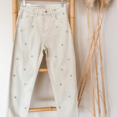 Calças bege claro com corações verdes penduradas num cabide de madeira sobre estrutura de bambu