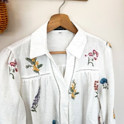Camisa branca com bordados florais coloridos pendurada em cabide de madeira