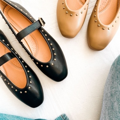 Dois pares de sapatos Mary Jane, um preto com fivela e tachas, e outro camel com tachas, sobre tecido branco, com jeans azul ao lado.