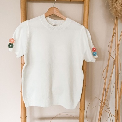 Camisola branca de malha com flores coloridas nas mangas pendurada em cabide de madeira.