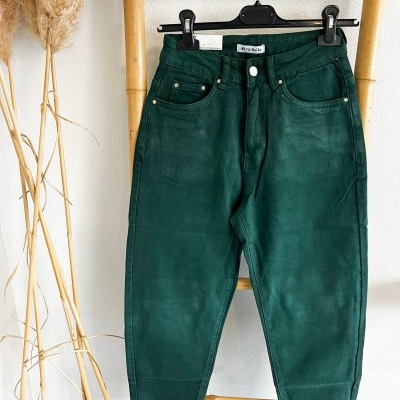 Calças verdes escuras penduradas numa escada de madeira com etiqueta 'Mara Made' e folhas secas ao lado.