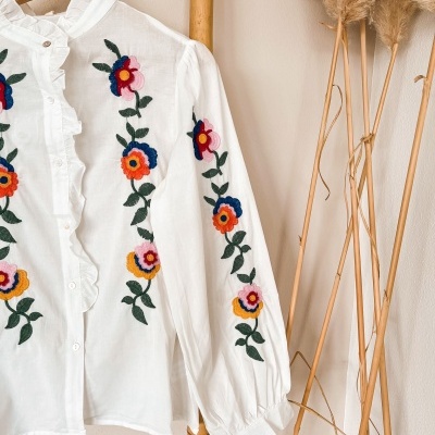 Camisa branca com bordados florais coloridos pendurada num cabide de madeira