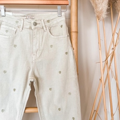 Calças de ganga brancas com corações verdes penduradas numa estrutura de bambu com plantas secas ao lado.