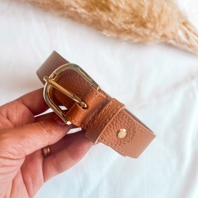 Cinto castanho em couro com fivela dourada segurado pela mão com fundo branco e planta decorativa