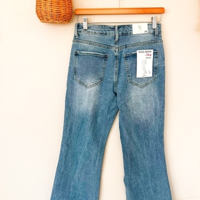 Calças de ganga azuis penduradas num cabide de madeira com etiqueta FOLY ROSE -5kg Jeans XS e cesto de vime com flores secas no suporte de parede