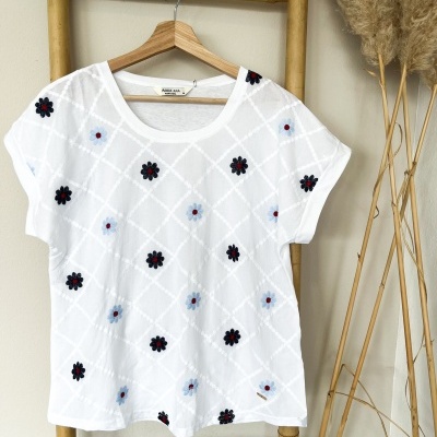 T-shirt branca com flores bordadas em azul e preto pendurada num cabide numa escada de bambu