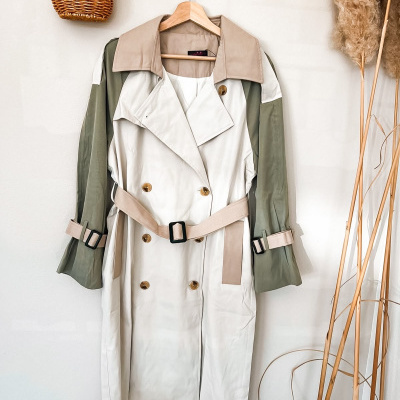 Casaco comprido tipo trench coat bege, verde e castanho num cabide de madeira numa parede clara.