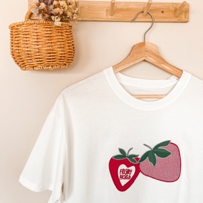 T-shirt branca com estampa de morangos pendurada num cabide de madeira num suporte de parede com cesta trançada.