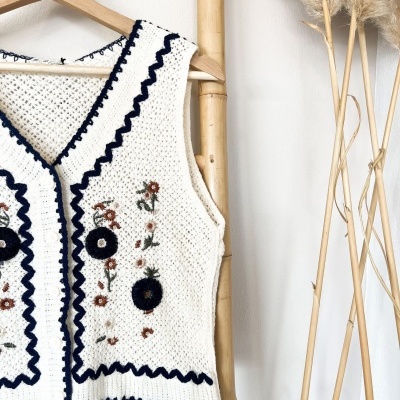 Colete de malha branco bordado com flores coloridas, pendurado em cabide de madeira sobre suporte de bambu com plantas decorativas ao lado.