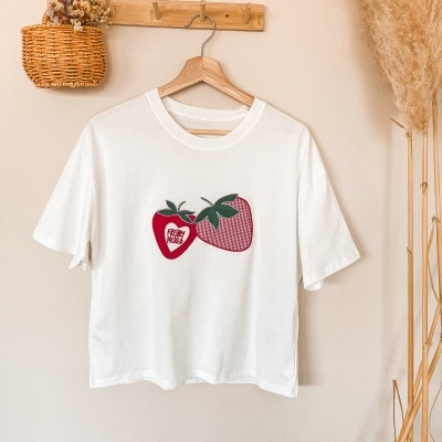 T-shirt branca com estampa de morangos e texto pendurada num cabide de madeira numa parede creme