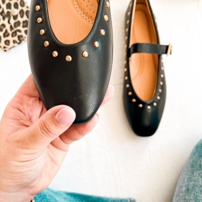 Sapatilhas pretas com tachas douradas e tira, interiores castanhos, em fundo branco e detalhe de peça azul e padrão animal print.