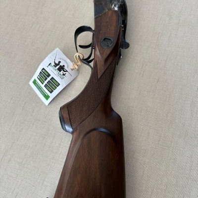 Espingarda com coronha em madeira castanha escura e detalhes em aço escuro sobre superfície bege.