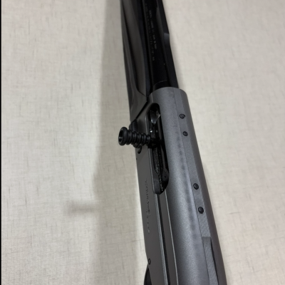 Espingarda preta com ação cinzenta e mola visível sobre fundo creme.