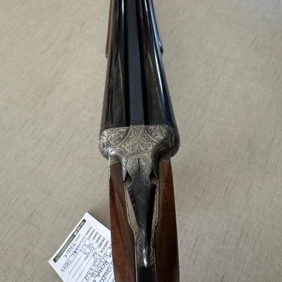 Espingarda de canos paralelos em metal preto com coronha em madeira castanha sobre superfície bege clara