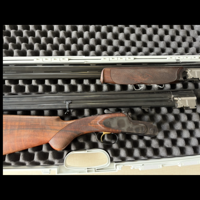 Espingarda desmonada com coronha de madeira e canos pretos numa caixa com interior de espuma preta acolchoada.
