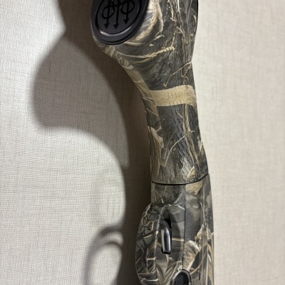 Espingarda de caça camuflada com fole e gatilho encostada em parede clara