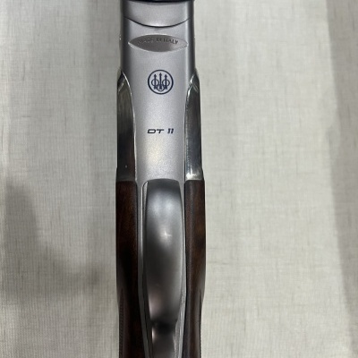 Parte superior de uma arma de caça com madeira escura e metal prateado, com texto e logo visíveis.