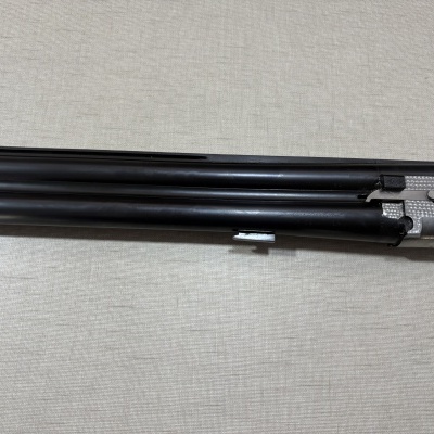 Parte de espingarda de canos duplos em metal preto e prateado sobre superfície cinzento claro.