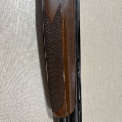 Espingarda com coronha de madeira castanha texturizada e canos pretos encostada à parede bege