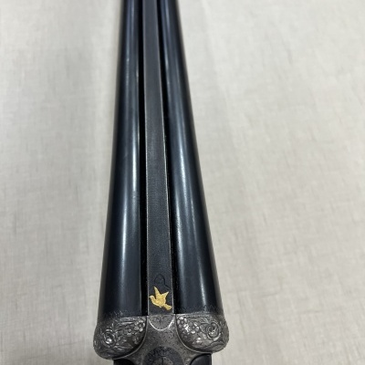 Espingarda de canos paralelos em metal preto com detalhes prateados e símbolo dourado sobre tecido bege