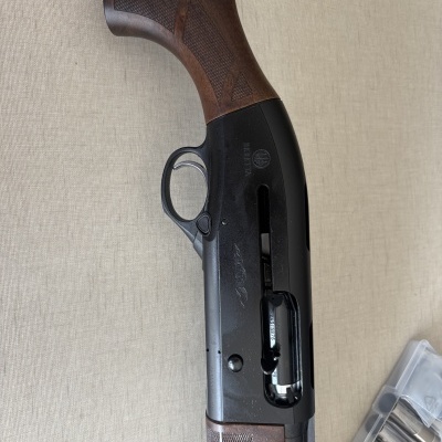 Espingarda com coronha de madeira e corpo metálico preto sobre superfície bege com embalagem ao lado