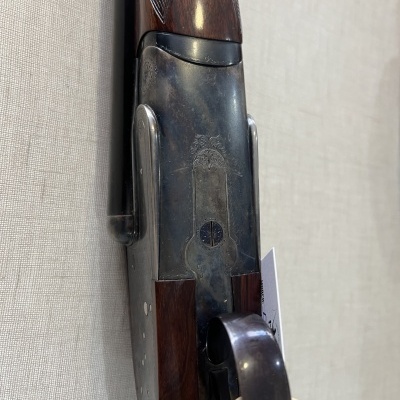 Espingarda com ação metal azulada e madeira escura, pendurada numa parede clara com etiqueta