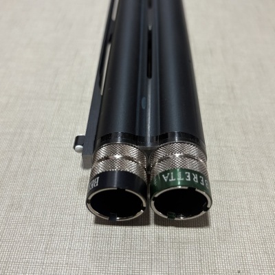 Detalhe dos canos traseiros de uma espingarda preta com inscrições BERETTA nas extremidades metálicas.