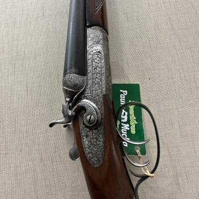 Espingarda de canos paralelos com madeira e metal entalhado, etiqueta verde com texto.