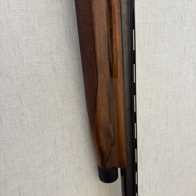 Parte de arma de caça com punho em madeira castanha polida e cano metálico negro encostada numa parede de tecido claro