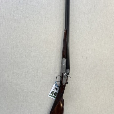 Espingarda de cano duplo com madeira castanha escura e metal preto pendurada em fundo cinza claro.