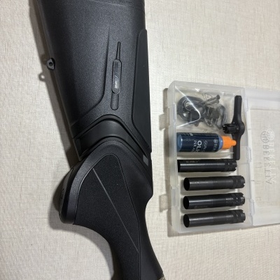 Coronha preta de espingarda Beretta ao lado de estojo transparente com chokes metálicos e acessórios.