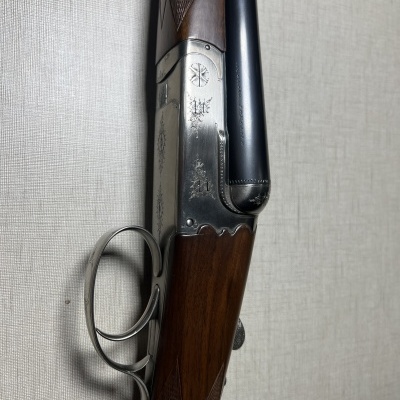 Espingarda dupla com madeira e metal, detalhes gravados, encostada numa parede cinza clara