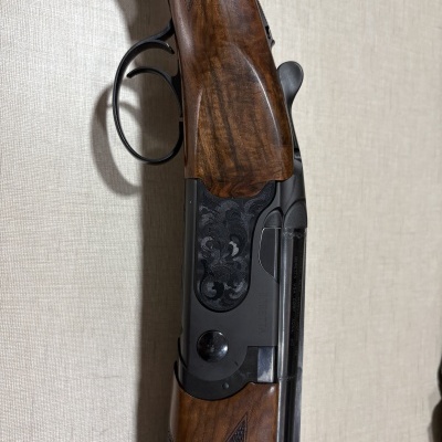 Parte próxima do gatilho e coronha de uma espingarda em madeira e metal preto com detalhes entalhados.