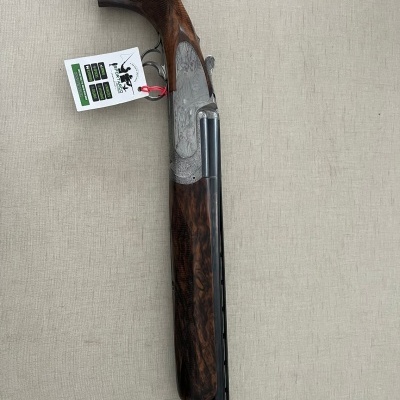 Espingarda com coronha em madeira escura e detalhes metálicos, etiqueta branca presa, sobre fundo bege.